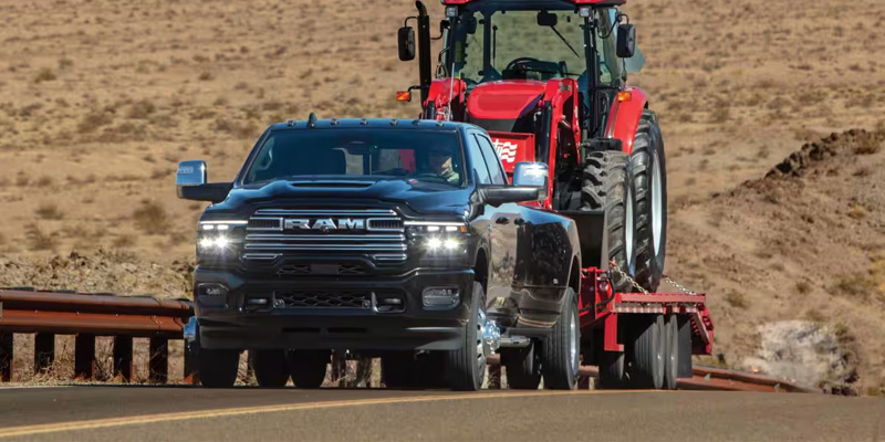 A 2026 RAM 3500 in Diamond Black Crystal Pearl-Coat towing a trailer and farm tractor, with optional diesel gas
